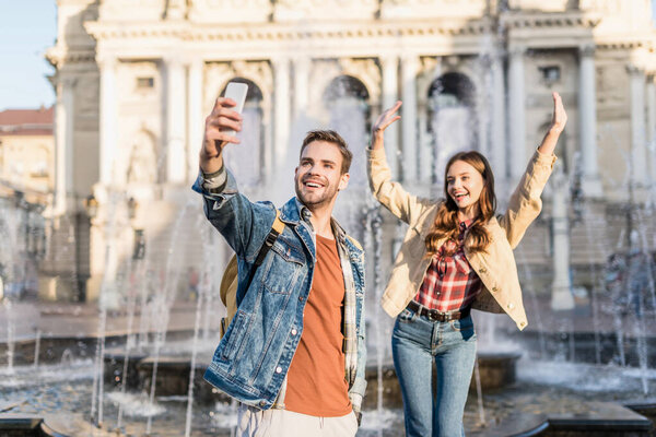 Man with girlfriend smiling and taking selfie with smartphone near fountain in city