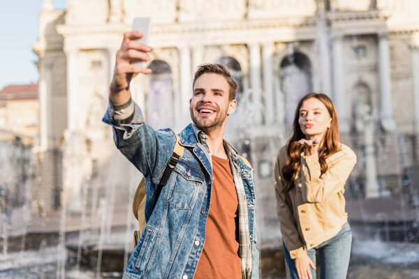 Selective focus of happy man with girlfriend blowing kiss taking selfie with smartphone near fountain in city