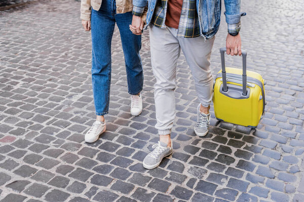 Cropped view of couple holding hands and traveling with suitcase together