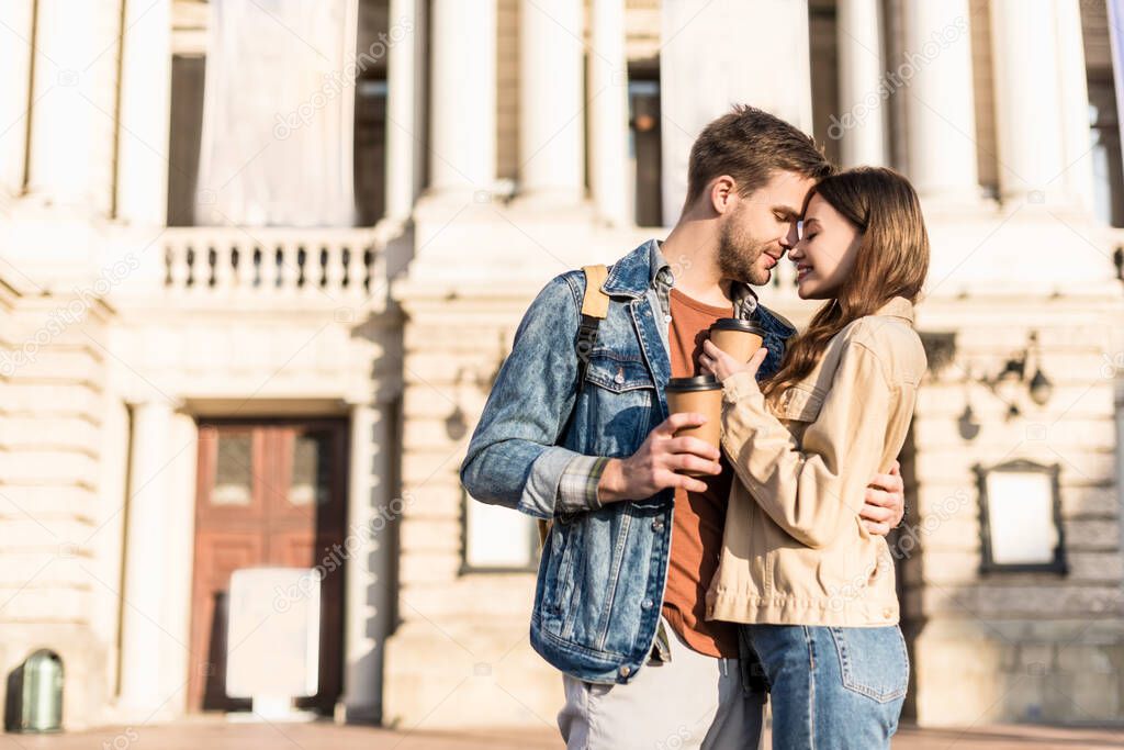 Boyfriend and girlfriend with disposable cups of coffee hugging in city