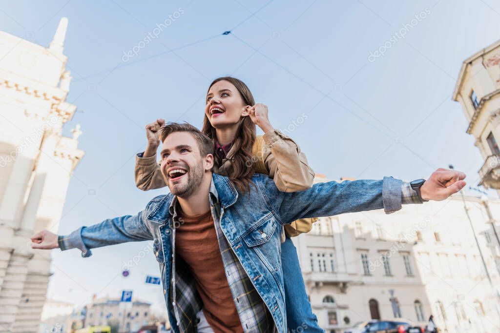 Low angle view of boyfriend smiling and piggybacking excited girl with open arms in city