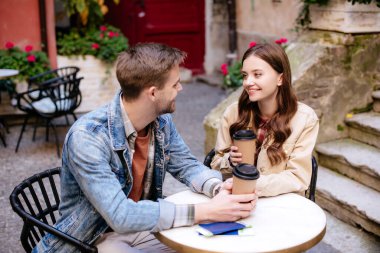 Selective focus of couple with disposable cups of coffee and passports looking at each other in cafe in city clipart