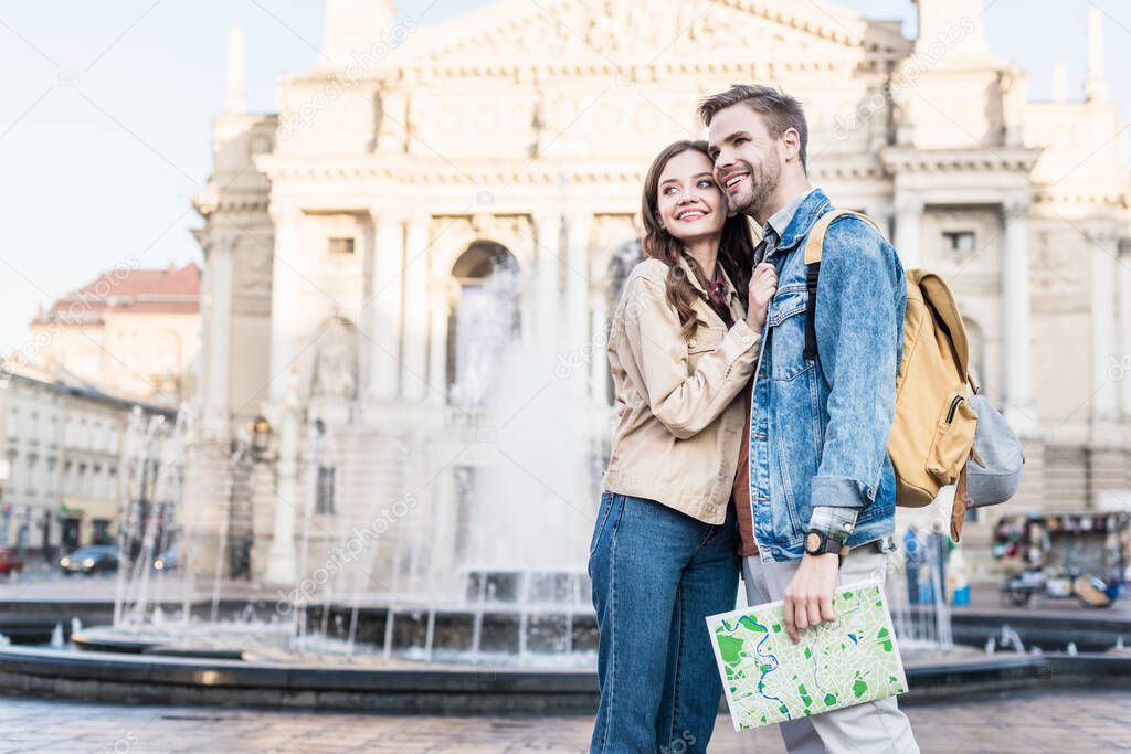 Couple looking away, hugging and smiling with map near fountain in city