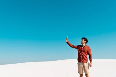 Kumsalda yakışıklı bir adam açık mavi gökyüzüne karşı akıllı telefon kullanıyor.