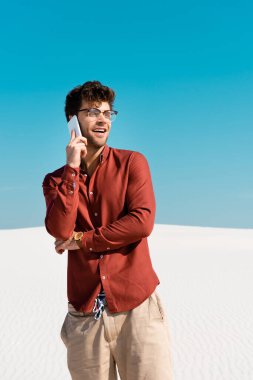 Kumsalda mutlu yakışıklı bir adam açık mavi gökyüzüne karşı akıllı telefonla konuşuyor.