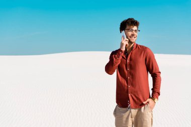 Kumsalda gülümseyen yakışıklı adam açık mavi gökyüzüne karşı akıllı telefondan konuşuyor.