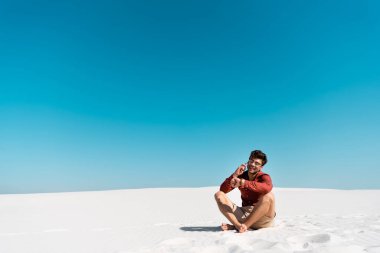 Kumsalda mutlu bir adam akıllı telefondan konuşuyor ve açık mavi gökyüzüne karşı kol saatine bakıyor.