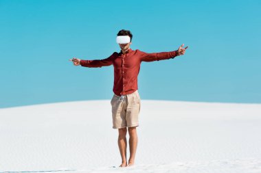 VR kulaklıklı, kumlu sahilde açık mavi gökyüzüne karşı kolları olan bir adam.