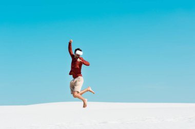 Kumlu sahilde, kulaklıklı bir adam açık mavi gökyüzüne doğru atlıyor.