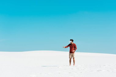 VR kulaklıklı kumsaldaki adam açık mavi gökyüzüne işaret ediyor.