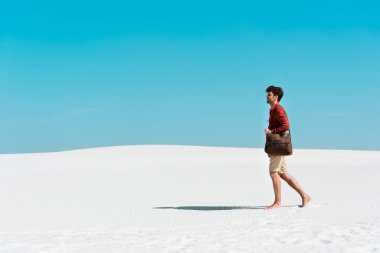 Deri çantalı yakışıklı adam kumlu sahilde açık mavi gökyüzüne doğru yürüyor.