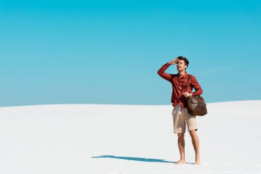 Deri çantalı yakışıklı adam kumlu sahilde açık mavi gökyüzüne bakıyor.