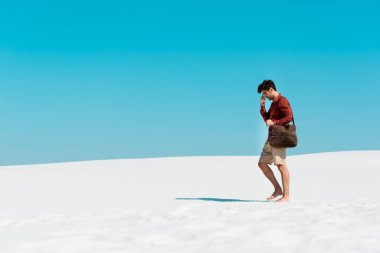 Deri çantalı yakışıklı adam kumlu sahilde açık mavi gökyüzüne doğru yürüyor.