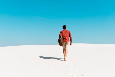 Deri çantalı bir adam kumlu sahilde açık mavi gökyüzüne doğru yürüyor.