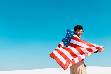 Rüzgarlı kumlu sahilde, açık mavi gökyüzüne karşı Amerikan bayrağı taşıyan mutlu adam.