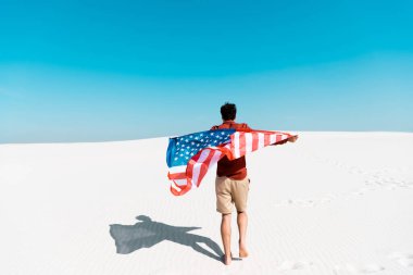 Rüzgarlı kumlu sahilde açık mavi gökyüzüne karşı Amerikan bayrağı taşıyan adamın arka görüntüsü