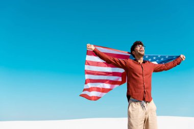 Rüzgarlı kumlu sahilde, açık mavi gökyüzüne karşı Amerikan bayrağı taşıyan mutlu adam.