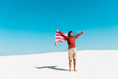 Rüzgarlı kumlu sahilde, açık mavi gökyüzüne karşı Amerikan bayrağı taşıyan mutlu adam.