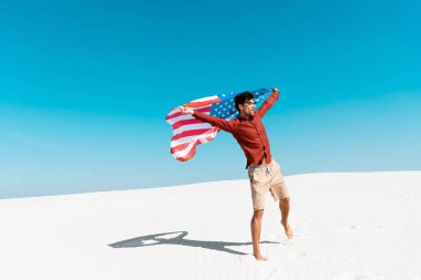 Rüzgarlı kumlu sahilde, açık mavi gökyüzüne karşı Amerikan bayrağı taşıyan adam.