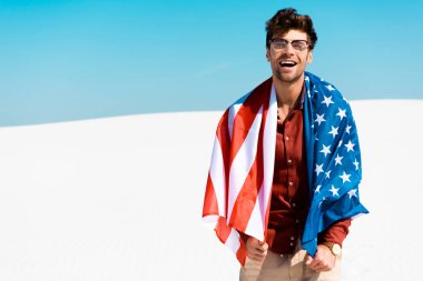 Kumlu sahilde, açık mavi gökyüzüne karşı Amerikan bayrağı taşıyan mutlu yakışıklı adam.