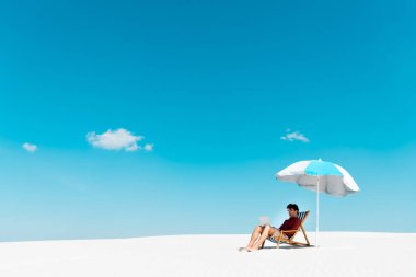 Kumlu sahilde mavi gökyüzüne karşı şemsiyenin altında dizüstü bilgisayarı olan serbest çalışan biri.