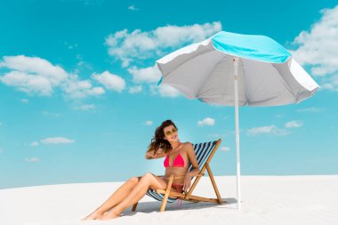 Gülümseyen, mayo giymiş, güneş gözlüklü, kumlu sahilde şemsiyenin altında oturan güzel seksi kız.