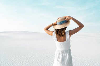 Beyaz elbiseli güzel bir kız ve kumlu sahilde mavi gökyüzü olan hasır bir şapka.