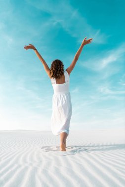 back view of beautiful girl in white dress with hands in air on sandy beach with blue sky clipart