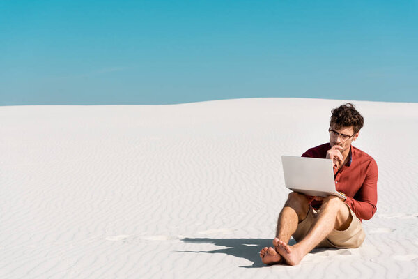 застенчивый красавец-фрилансер на песчаном пляже с ноутбуком на фоне голубого неба

