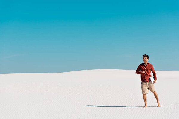 handsome freelancer on sandy beach with laptop against clear blue sky