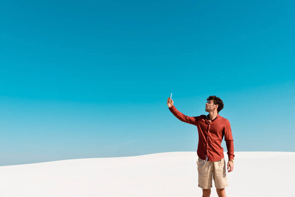 handsome man on sandy beach using smartphone against clear blue sky