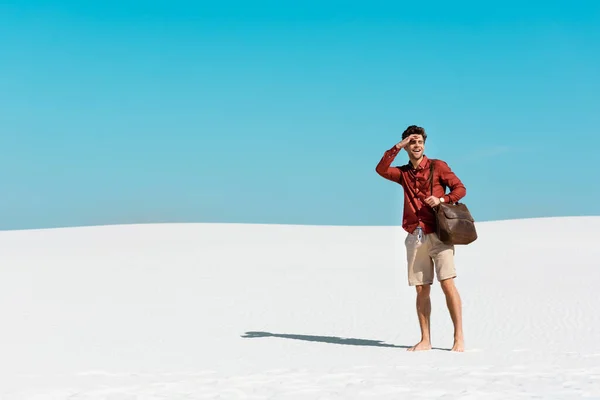 handsome man with leather bag  looking away on sandy beach against clear blue sky