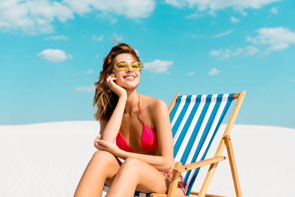 улыбающаяся красивая сексуальная девушка в купальнике и солнцезащитных очках, сидящая в шезлонге на песчаном пляже с голубым небом и облаками
