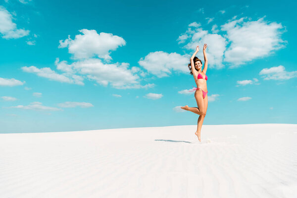 счастливая красивая сексуальная девушка в купальнике, прыгающая с руками в воздухе на песчаном пляже с голубым небом и облаками

