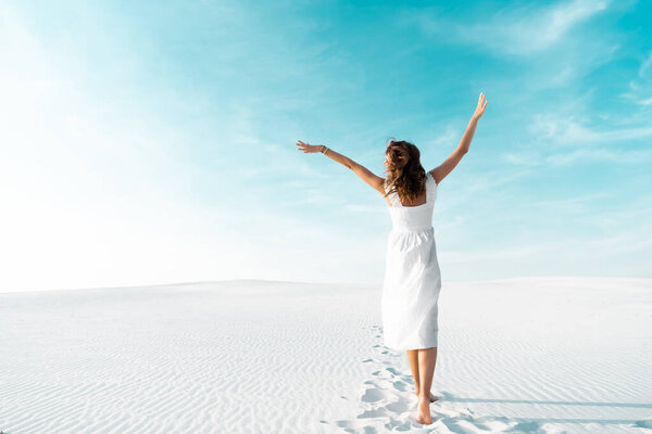 back view of beautiful girl in white dress with hands in air on sandy beach with blue sky
