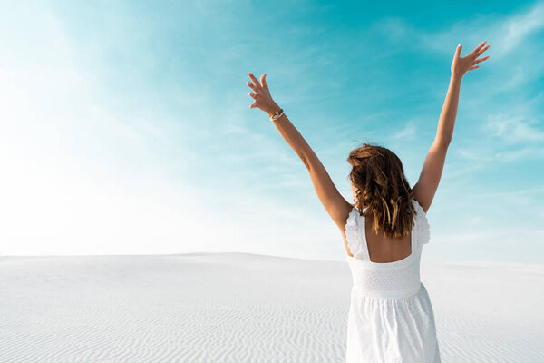 Вид сзади красивой девушки в белом платье с руками в воздухе на песчаном пляже с голубым небом
