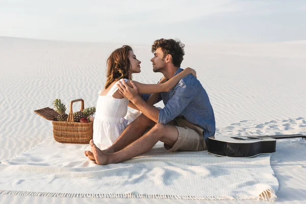 一对年轻夫妇抱在怀里 坐在毛毯上 沙滩上挂着一筐水果和声吉他 — 图库照片
