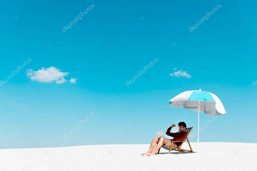 freelancer sitting with laptop in deck chair under umbrella on sandy beach against blue sky