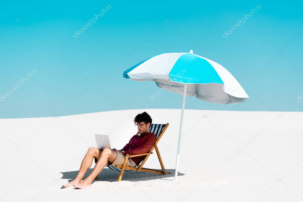 freelancer sitting with laptop in deck chair under umbrella on sandy beach against blue sky