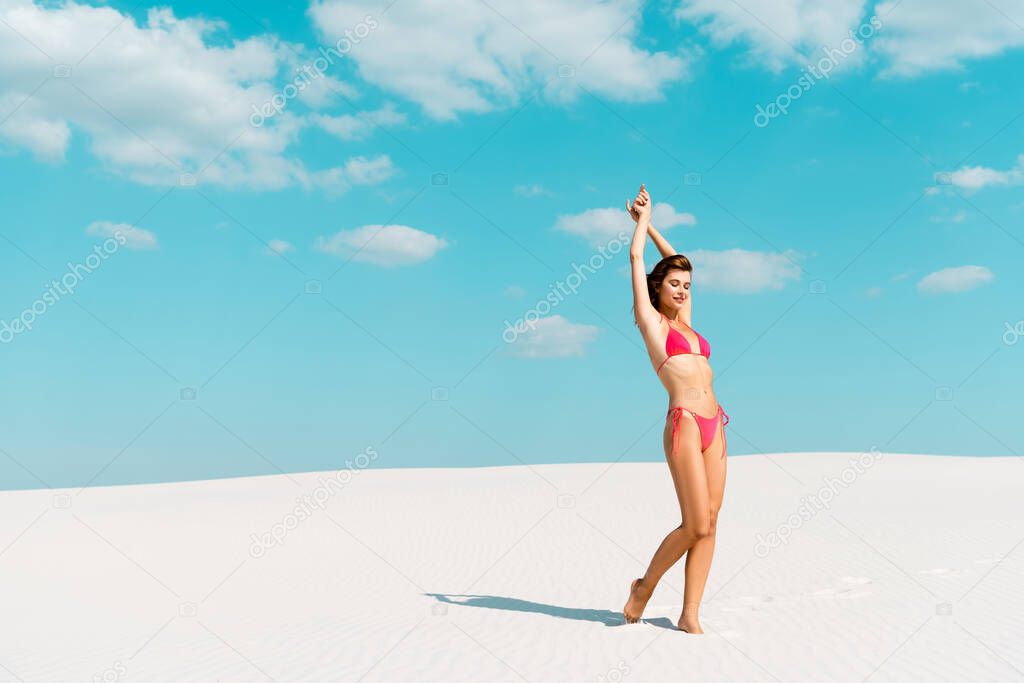 beautiful sexy girl in swimsuit with hands in air on sandy beach with blue sky and clouds