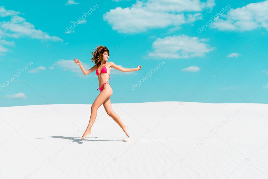 beautiful sexy girl in swimsuit jumping on sandy beach with blue sky and clouds