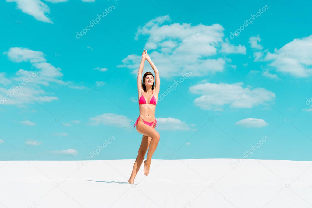 smiling beautiful sexy girl in swimsuit jumping with hands in air on sandy beach with blue sky and clouds