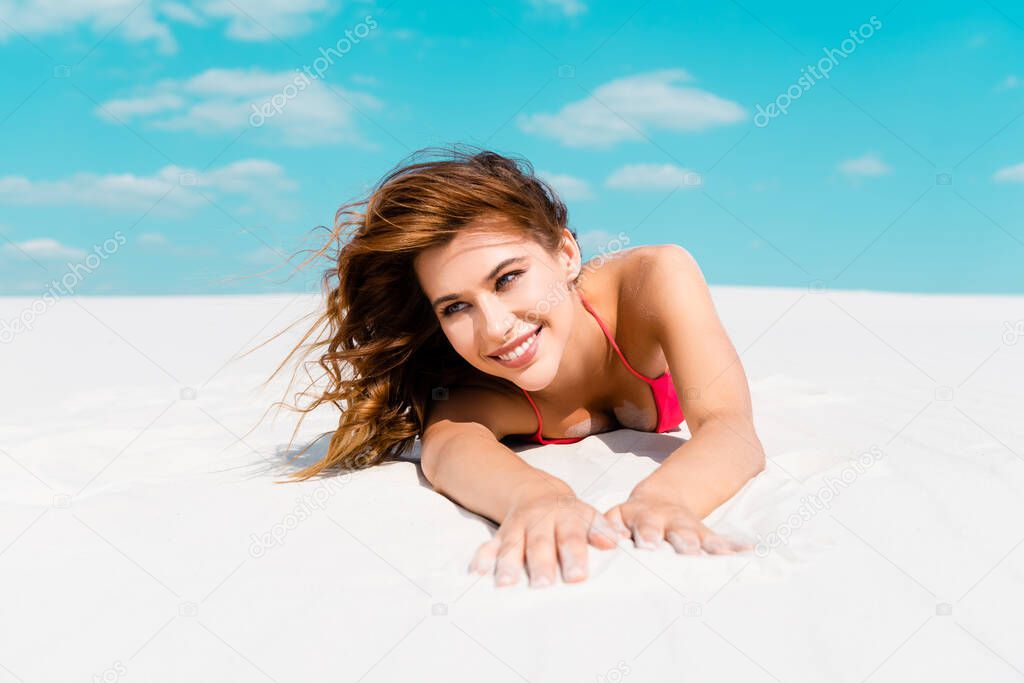 smiling beautiful sexy girl in swimsuit lying on sandy beach with blue sky and clouds at background