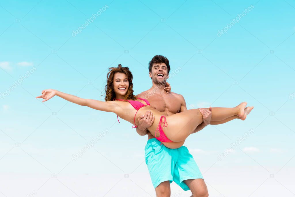 low angle view of young man holding beautiful sexy girlfriend in arms on sandy beach