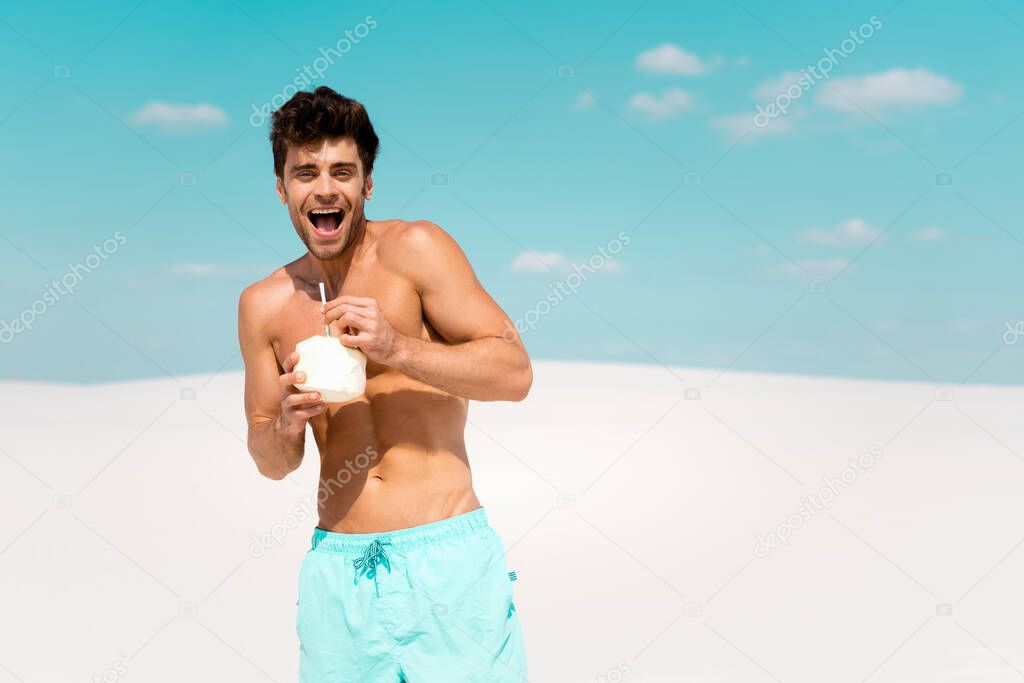 happy sexy man with muscular torso in swim shorts with coconut drink on sandy beach