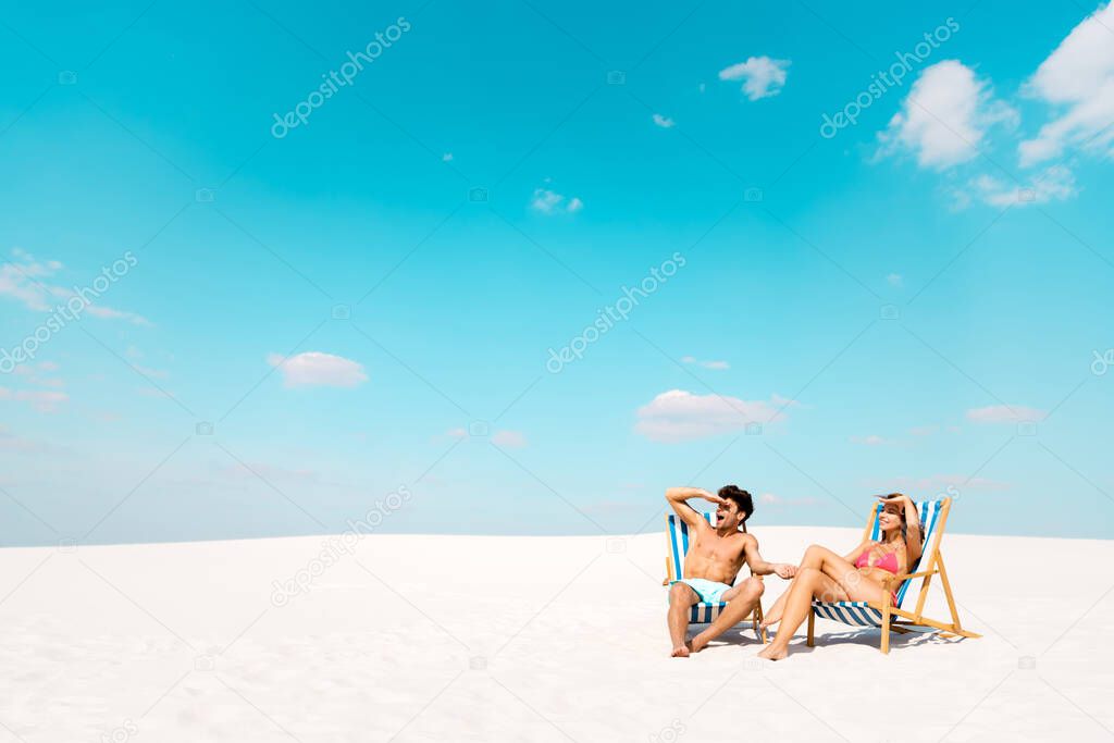 smiling young couple sitting in deck chairs and looking away on sandy beach
