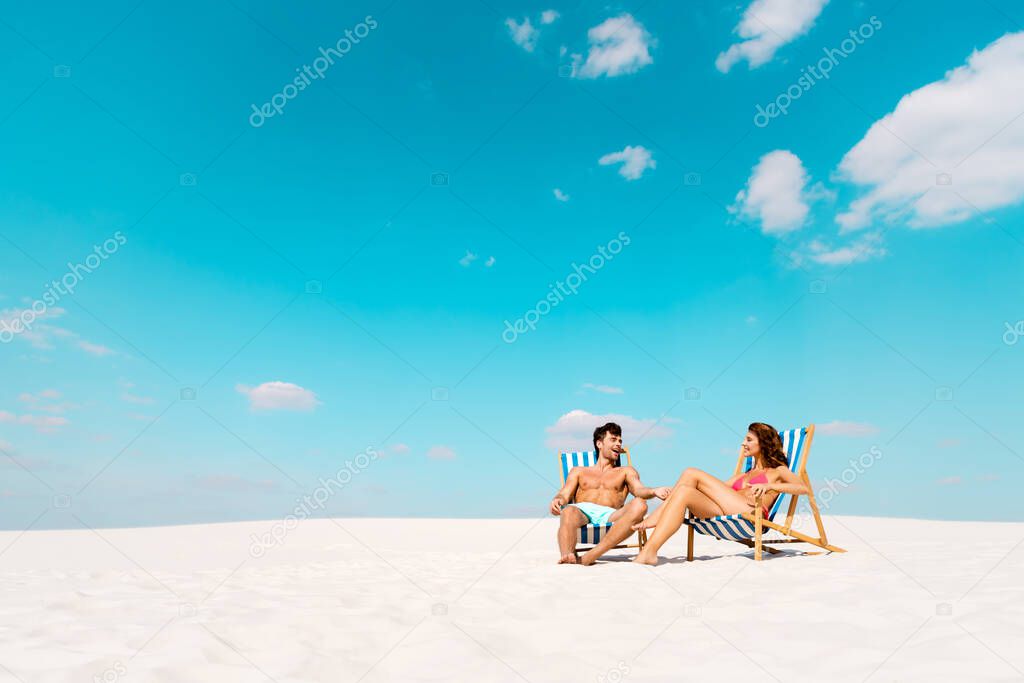 smiling young couple sitting in deck chairs on sandy beach