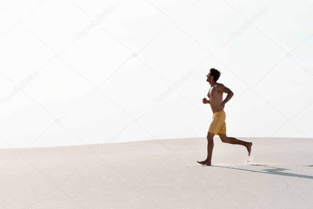man in swim shorts with muscular torso running on sandy beach