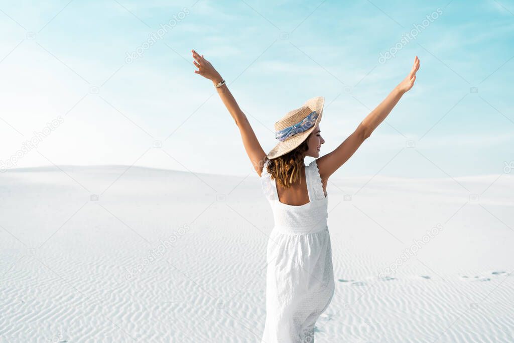 back view of beautiful girl in white dress and straw hat on sandy beach with blue sky