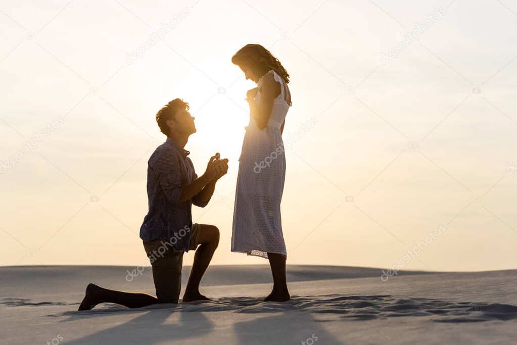 side view of young man doing marriage proposal to girlfriend on sandy beach at sunset